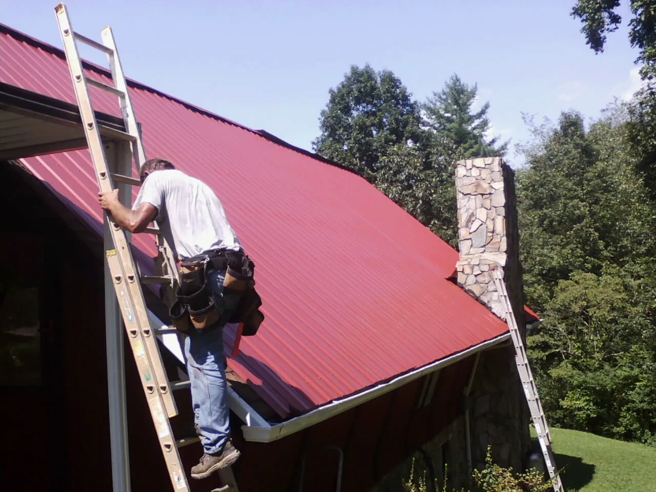 Roofer on a ladder beside a red metal roof and stone chimney — Soffit & Fascia Repair by Roofing Brothers in Holloway